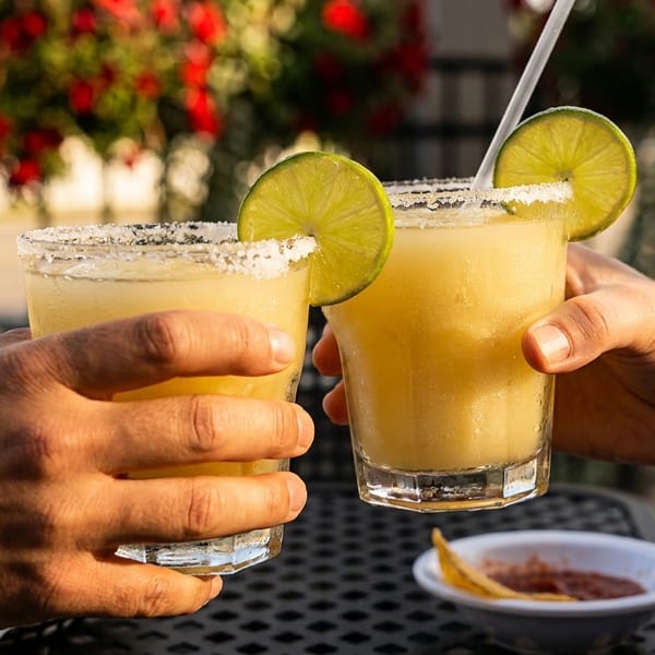 Two hands clinking frozen margaritas with lime wedges and salt rims on an outdoor patio.