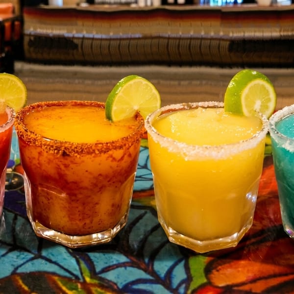 Four colorful frozen margaritas, red, orange, yellow, and blue, with lime and rim garnishes.