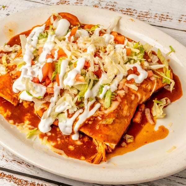 Two enchiladas covered in red sauce, topped with lettuce, tomato, shredded cheese, and sour cream on a white plate.
