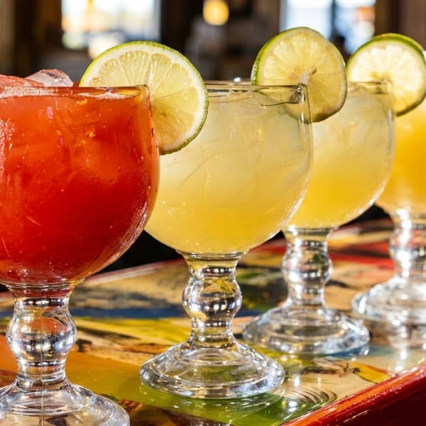 Four vibrant margaritas, one red and three yellow, garnished with lime slices on a colorful table.