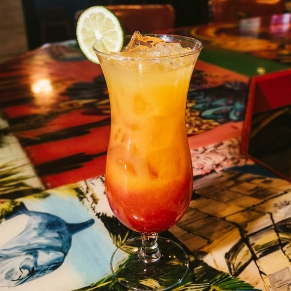 Tall glass with a layered orange and red cocktail, garnished with a lime slice on a colorful bar.