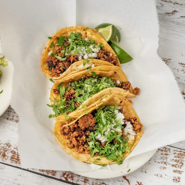 Three street tacos with meat, cilantro, and onions, served with lime and a side of guacamole.