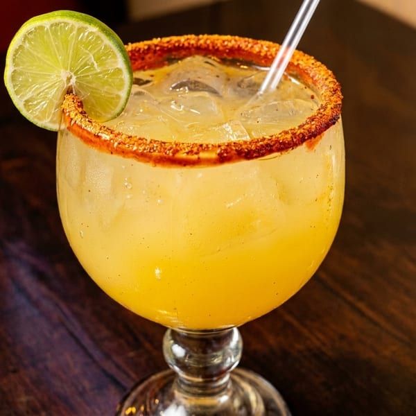 Yellow margarita with ice, a chili-salted rim, and a fresh lime wedge on a wooden table.